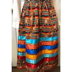 K&ouml;nnte beinhalten: Langer, brauner Rock mit s&uuml;dwestlichem Muster und farbenfrohem Bandbesatz. Der Rock hat einen breiten G&uuml;rtel mit drei silberfarbenen Schnallen. Die B&auml;nder sind in Blau-, Orange-, Gelb- und Rott&ouml;nen gehalten.