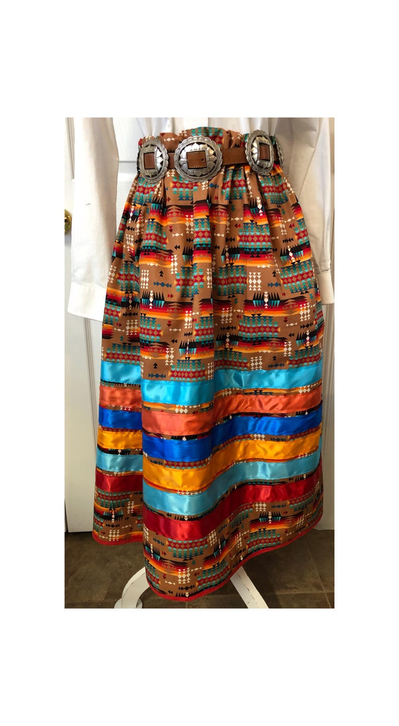 K&ouml;nnte beinhalten: Langer, brauner Rock mit s&uuml;dwestlichem Muster und farbenfrohem Bandbesatz. Der Rock hat einen breiten G&uuml;rtel mit drei silberfarbenen Schnallen. Die B&auml;nder sind in Blau-, Orange-, Gelb- und Rott&ouml;nen gehalten.