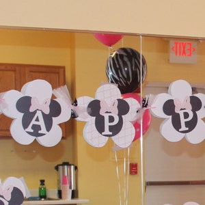 May include: A pink, white, and black banner with the word "HAPPY" spelled out in white letters with black outlines. The banner is decorated with black and white Mickey Mouse ears and pink bows. The banner is hanging from the ceiling in a room with a mirror and balloons.
