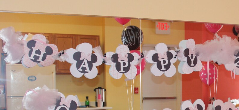 May include: A pink, white, and black banner with the word "HAPPY" spelled out in white letters with black outlines. The banner is decorated with black and white Mickey Mouse ears and pink bows. The banner is hanging from the ceiling in a room with a mirror and balloons.