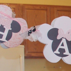 May include: A white and pink banner with the letters "E" and "A" in black and white. The letters are surrounded by black and white mouse ears and pink and white bows. The banner is decorated with white tulle and a black and white bow.