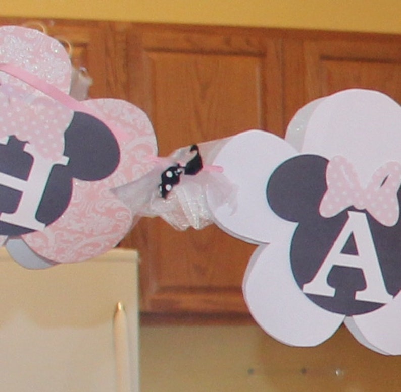May include: A white and pink banner with the letters "E" and "A" in black and white. The letters are surrounded by black and white mouse ears and pink and white bows. The banner is decorated with white tulle and a black and white bow.
