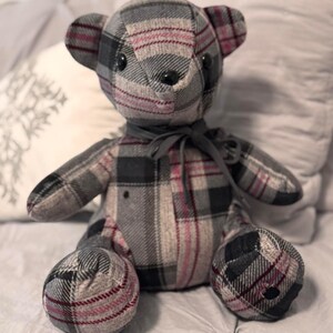 May include: A handmade teddy bear with a plaid pattern in shades of gray, black, and burgundy. The bear has black button eyes and a gray ribbon around its neck. The bear is sitting on a bed with pillows.