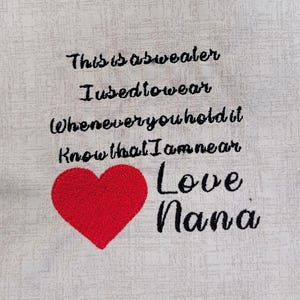 Könnte beinhalten: Ein weißer Stoff mit schwarzem gesticktem Text, der "This is a sweater I used to wear whenever you hold it know that I am near Love Nana" lautet, und einem roten gestickten Herzen.