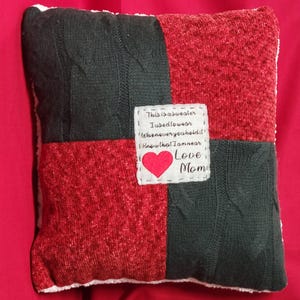 May include: A square throw pillow featuring a patchwork design of dark gray cable-knit fabric and textured red fabric. A white fabric patch in the center displays the embroidered text: This is a sweater I used to wear. Whenever you hold it, know that I am near. Love Mom, with a red heart.