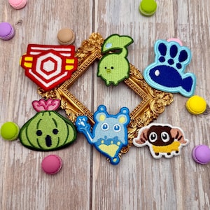 May include: A collection of six embroidered patches featuring colorful cartoon characters and shapes, including a red geometric emblem, a green creature, a blue fish-like figure, a green onion-shaped character, a blue mouse-like figure, and a ram.