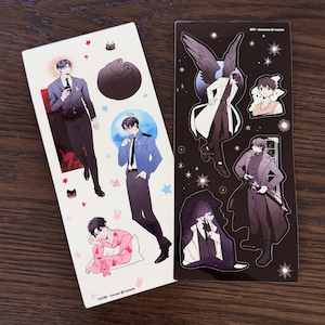 Puede incluir: Dos hojas de pegatinas con ilustraciones de estilo anime de personajes masculinos. Una hoja es blanca con personajes con trajes y ropa casual. La otra es negra con personajes con abrigos y alas de ángel. Incluye estrellas decorativas.