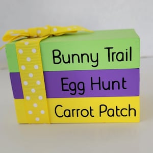 May include: A stack of three wooden blocks with a yellow ribbon with white polka dots. The blocks are painted in different colors: green, purple, and yellow. The text on the blocks reads "Bunny Trail", "Egg Hunt", and "Carrot Patch".