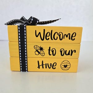 May include: A decorative yellow wooden block sign with the words "Welcome to our Hive" in black script. The sign features bee and honeycomb illustrations. A black and white ribbon is tied around the blocks.