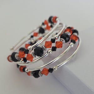 May include: A multi-strand wrap bracelet featuring alternating black and orange faceted crystal beads interspersed with small silver-toned metal spacer beads. The design consists of several coiled loops of silver-toned wire that create a layered appearance.
