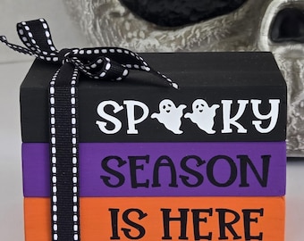 May include: A stack of three wooden blocks painted black, purple, and orange. The top block reads "SP00KY" with ghost graphics, the middle block says "SEASON", and the bottom block says "IS HERE". A black and white ribbon is tied around the blocks.