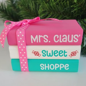 May include: Stacked decorative blocks in pink, white, and teal, tied with a pink polka dot ribbon. The top block reads "Mrs. Claus", the middle block says "Sweet" with candy graphics, and the bottom block says "Shoppe".