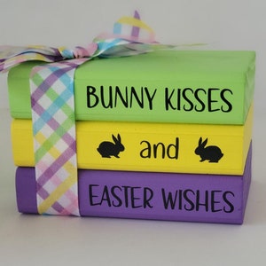 May include: A stack of three wooden blocks painted in green, yellow, and purple. The top block reads "Bunny Kisses", the middle block reads "and" with a black bunny silhouette on each side, and the bottom block reads "Easter Wishes". The blocks are tied together with a pink and blue plaid ribbon.