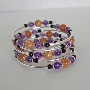 May include: A multi-strand coil bracelet featuring silver-tone wire wrapped in a spiral pattern. The bracelet is adorned with alternating round crackle glass beads in shades of purple and orange, accented by small, smooth black spherical beads.