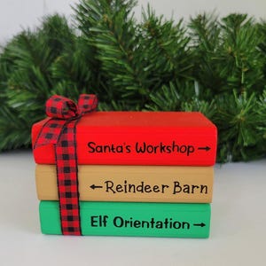 May include: A stack of three wooden books painted in red, tan, and green. The books are decorated with black and red plaid ribbon and have the words "Santa's Workshop", "Reindeer Barn", and "Elf Orientation" written on them.