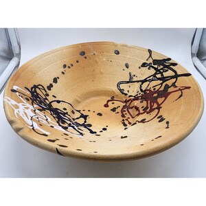 May include: A large, round, ceramic bowl with a light brown glaze. The bowl has a wide, shallow rim and is decorated with black, white, and red paint splatters and lines.