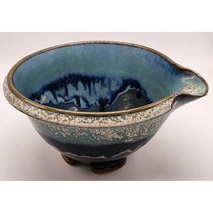May include: A handmade ceramic bowl with a pouring spout. The bowl features a gradient of blue and teal hues, with a speckled white and brown rim. The bowl is supported by three small feet.