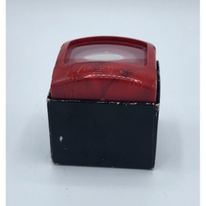 May include: A red plastic candle holder with a white candle inside. The candle holder has a black box with the words "Handy Stove" printed on it.