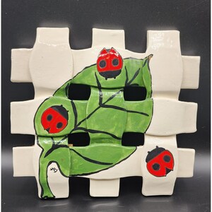 May include: A ceramic wall hanging with a woven design in off-white. A green leaf with three red and black ladybugs is centered on the piece. The ladybugs have black spots and the leaf has black outlines.