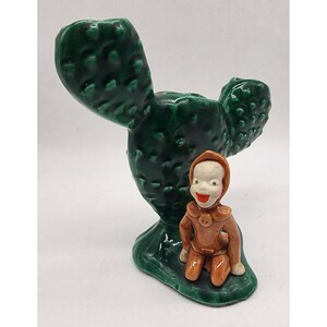 May include: A green ceramic cactus figurine with a small brown ceramic figure sitting at its base. The figure has a smiling face and is wearing a brown outfit.