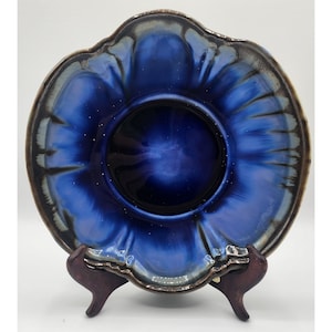 May include: A blue and brown ceramic plate with a scalloped edge. The plate has a deep blue glaze with a subtle iridescent sheen. The plate is on a wooden stand.