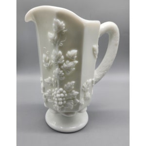 May include: White milk glass pitcher with a grapevine design. The pitcher has a wide mouth and a handle. The base of the pitcher is round.