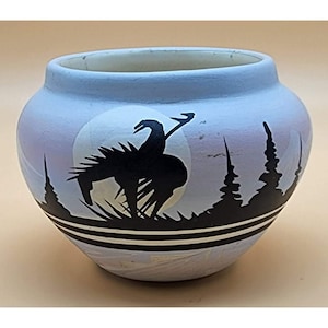 May include: A blue ceramic pot with a black silhouette of a Native American on horseback riding into a full moon. The pot has a black and white band around the rim.