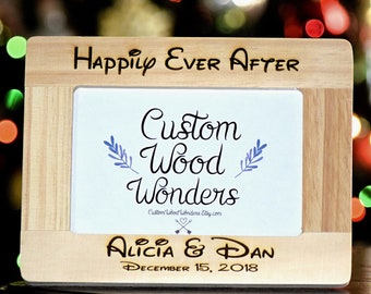 May include: A light wood picture frame with the words "Happily Ever After" engraved on the top. The frame has a white insert with the words "Custom Wood Wonders" and a small blue floral design. The bottom of the frame has the names "Alicia & Dan" and the date "December 15, 2018" engraved in a gold color.