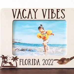 Może przedstawiać: Biała drewniana ramka na zdjęcia z tekstem "VACAY VIBES" u góry i "FLORIDA 2022" na dole. Ramka ma wycięcie palm i żółwia morskiego. Ramka zawiera zdjęcie dziecka w żółtej kamizelce ratunkowej stojącego w oceanie.