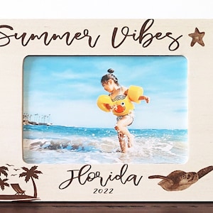 Op de afbeelding: Een wit houten fotolijstje met de tekst "Summer Vibes" en een stervormig ontwerp bovenaan. Het lijstje bevat een foto van een kind in een gele zwemvest dat in de zee speelt. Onderaan het lijstje staat de tekst "Florida 2022" met palmbomen en een zeeschildpad-ontwerp.