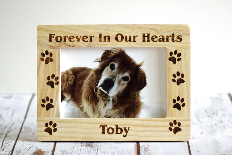 May include: Wooden photo frame with a brown dog inside. The frame has the words "Forever In Our Hearts" and "Toby" engraved on it. The frame is decorated with paw prints.