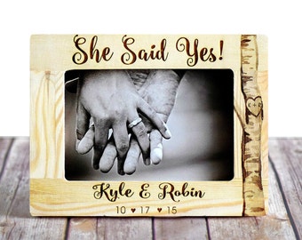 May include: A wooden picture frame with a birch tree design. The frame has a photo of a couple holding hands with an engagement ring on the woman's finger. The frame is engraved with the text "She Said Yes!" and "Kyle & Robin 10 ♥ 17 ♥ 15".