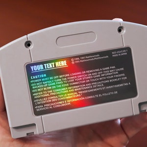 Op de afbeelding: Een grijze Nintendo 64-gamecartridge met een holografische sticker waarop "YOUR TEXT HERE" staat. De sticker bevat ook een lijst met veiligheidsvoorzorgsmaatregelen in het Engels, Frans en Spaans.