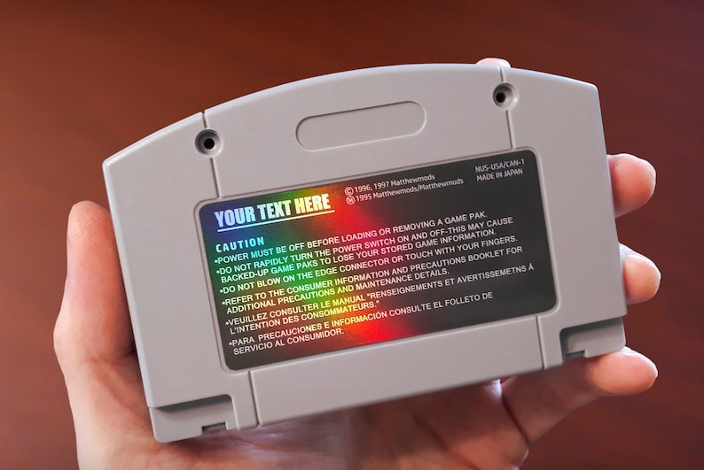 Op de afbeelding: Een grijze Nintendo 64-gamecartridge met een holografische sticker waarop "YOUR TEXT HERE" staat. De sticker bevat ook een lijst met veiligheidsvoorzorgsmaatregelen in het Engels, Frans en Spaans.