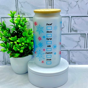 May include: A white frosted glass tumbler with a wooden lid and a clear straw. The tumbler has a snowflake design and the words "Decaf", "Shots", "Syrup", "Milk", "Custom", and "Drink" with checkboxes next to each.