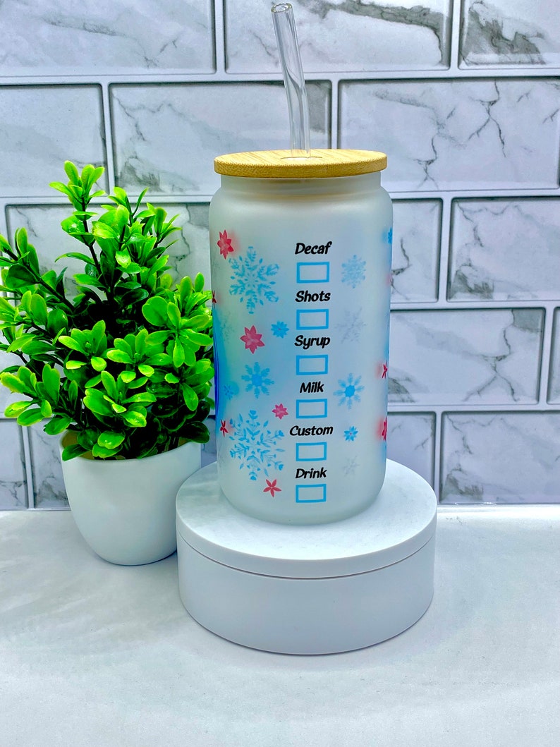May include: A white frosted glass tumbler with a wooden lid and a clear straw. The tumbler has a snowflake design and the words "Decaf", "Shots", "Syrup", "Milk", "Custom", and "Drink" with checkboxes next to each.
