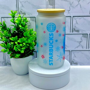 May include: A frosted glass Starbucks tumbler with a blue and white snowflake design. The tumbler has a bamboo lid and a clear plastic straw. The Starbucks logo is visible on the front of the tumbler.