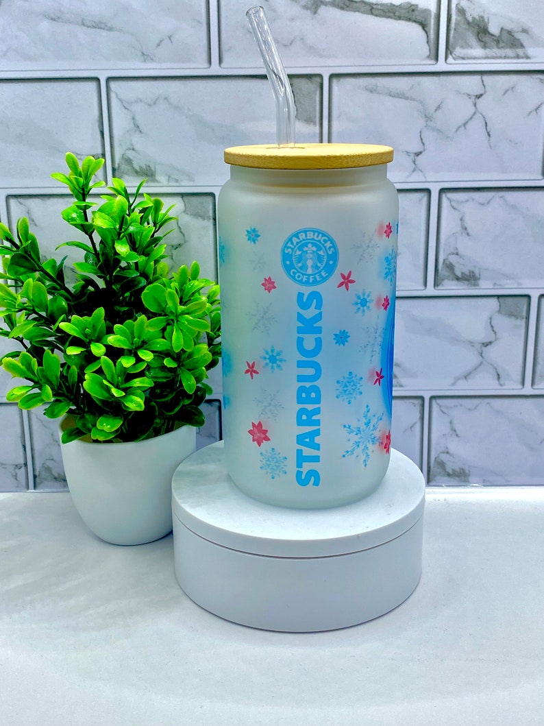 May include: A frosted glass Starbucks tumbler with a blue and white snowflake design. The tumbler has a bamboo lid and a clear plastic straw. The Starbucks logo is visible on the front of the tumbler.