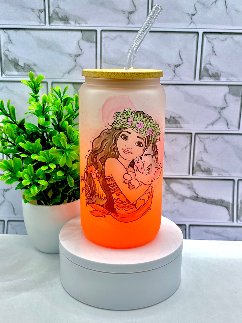 May include: A frosted glass water bottle with a bamboo lid and a straw. The bottle features a colorful illustration of a cartoon character with a pig. The bottle is on a white pedestal.