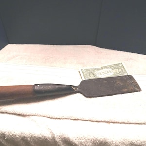 May include: A metal wood chisel with a dark brown wooden handle and a flat, rectangular steel blade. The tool is positioned next to a one-dollar bill for scale, showing the blade width relative to the currency. The metal shows signs of age and wear.