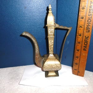 May include: A small, ornate brass teapot with intricate floral designs. The teapot has a tall, slender spout and a rounded body. The base of the teapot is a square shape.