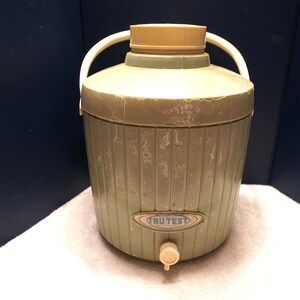 May include: Vintage olive green and tan water cooler with a spigot and white handle. The cooler has a ribbed design and a tan lid. The label reads "TRU-TEST".