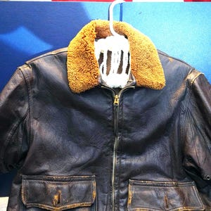 May include: A vintage brown leather jacket with a tan shearling collar, hanging on a white hanger. The jacket has a zipper closure, two front pockets, and a distressed appearance, suggesting age and wear. The jacket is a classic outerwear piece.