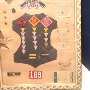 May include: A framed display featuring a black vest-shaped fabric piece adorned with various embroidered patches, including three diamond-shaped Cub Scout badges, rows of arrow-shaped pins, and a patch labeled Taylor Texas, alongside several paper documents.