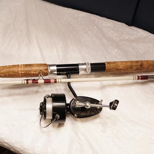 May include: A fishing rod with a white shaft, cork handle, and maroon accents, paired with a black spinning reel. The rod features silver metal hardware and a white line visible on the reel spool. The assembly is presented in a horizontal orientation.