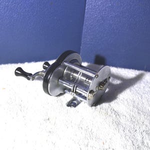 Puede incluir: Un carrete de pesca vintage con cuerpo plateado y mango negro. El carrete tiene un diseño circular con múltiples componentes y una manivela. El carrete está diseñado para la pesca en agua salada.