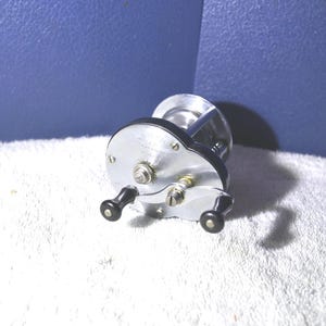 May include: A vintage fishing reel with a silver body and black handles. The reel has a circular design with several screws and a winding mechanism. The reel is set against a blue background and a white, textured surface.