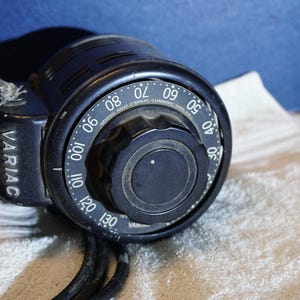 May include: A black Variac autotransformer with a dial displaying numbers from 0 to 130. The dial has a prominent knob for adjusting voltage. The device is on a white cloth with a blue background.