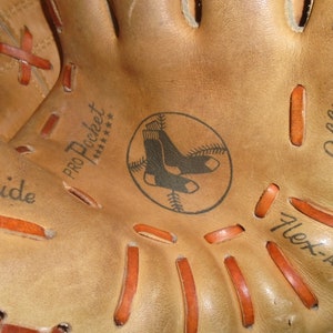 May include: Close-up of a tan leather baseball glove with orange stitching. The glove has the words "Cowhide", "Pro Pocket", and "Flex-Action" printed on it. A circular logo with two black socks inside a baseball is also visible.