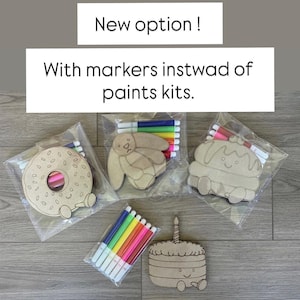 May include: Three clear plastic bags containing light brown wooden cutouts in the shapes of a donut, a rabbit, and a cupcake, each paired with a set of colorful markers. A separate pack of markers and a wooden cake cutout are also visible. Text reads: New option! With markers instwad of paints kits.